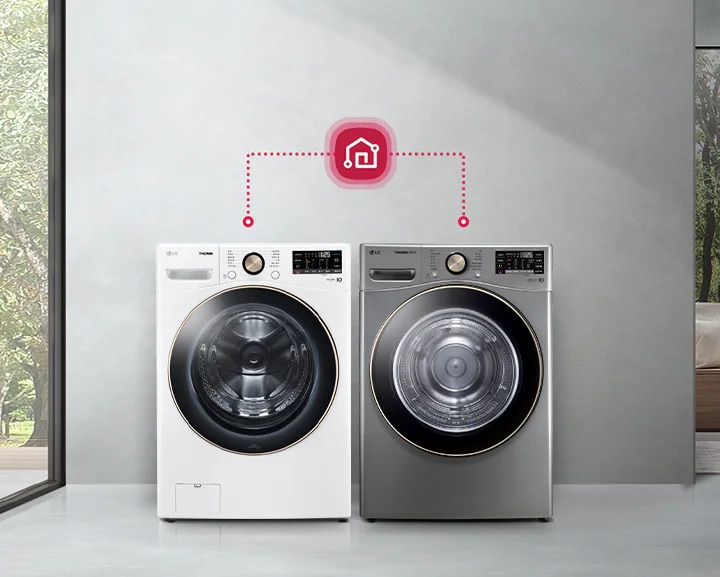 두 대의 세탁기가 나란히 놓여 있으며, 각각 흰색과 회색으로 되어 있습니다. 세탁기 위에는 집 모양의 아이콘이 연결되어 있어 스마트 기능을 암시합니다. 주변은 넓은 창문을 통해 보이는 나무와 자연 풍경으로 꾸며져 있으며, 실내는 현대적인 디자인의 공간입니다.