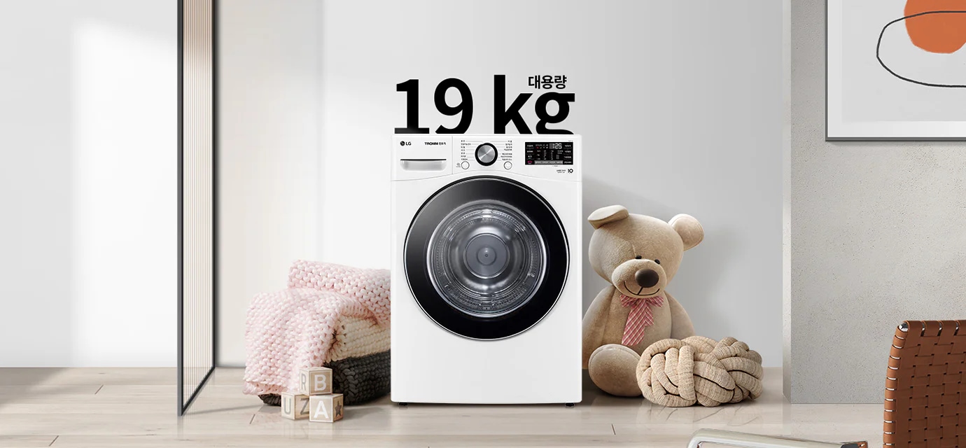 세탁기가 놓여 있으며, 상단에 19kg 대용량이라는 문구가 있습니다. 세탁기 옆에는 곰 인형과 두꺼운 니트 담요가 놓여 있고, 바닥에는 알파벳 블록이 있습니다. 벽에는 추상적인 그림이 걸려 있습니다.