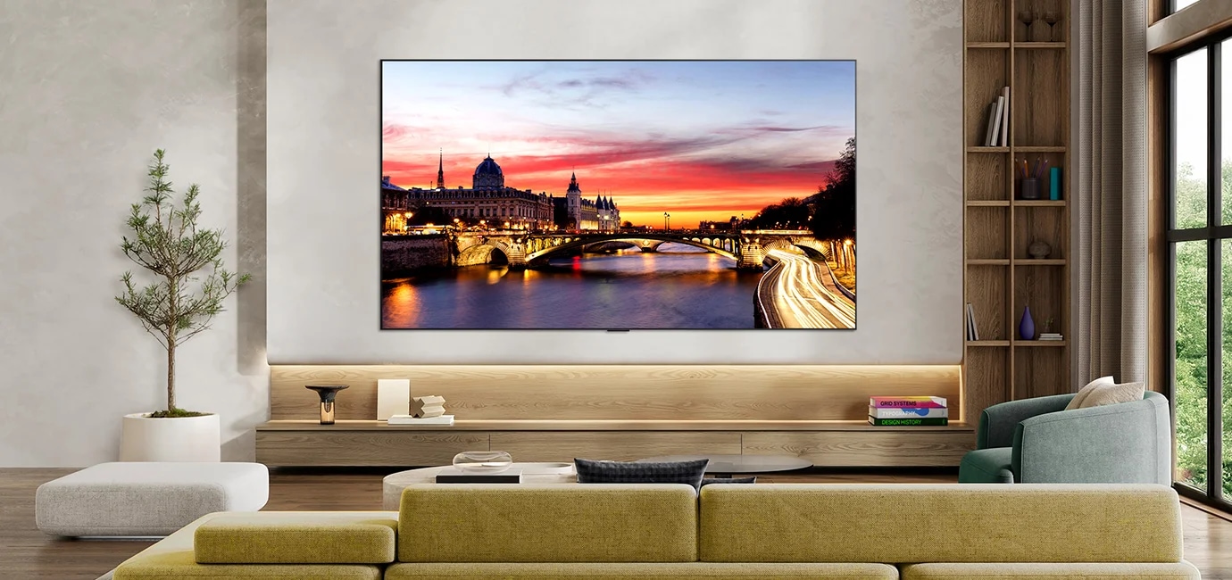 세련된 거실 공간에 벽걸이로 설치되어 있으며, 초슬림 디자인과 얇은 베젤을 갖추고 있습니다. 화면에는 해질녘 강가 도시 풍경이 표시되고 있습니다.