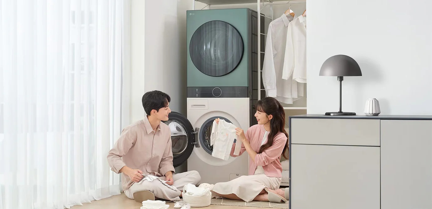 남성과 여성이 세탁기 앞에서 빨래를 정리하며 대화하고 있습니다. 남성은 베이지색 셔츠와 바지를, 여성은 분홍색 상의와 흰색 치마를 입고 있습니다. 세탁기는 두 대가 위아래로 놓여 있으며, 뒤쪽에는 옷걸이에 셔츠가 걸려 있습니다. 바닥에는 빨래 바구니와 접힌 옷들이 놓여 있고, 옆에는 램프가 있는 회색 가구가 보입니다. 창문에는 흰색 커튼이 드리워져 있습니다.