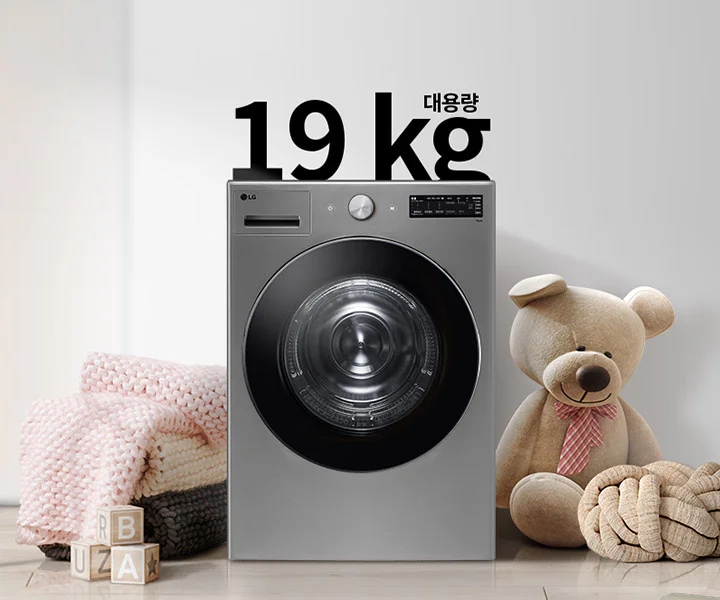 세탁기가 놓여 있으며, 그 위에 19kg 대용량이라는 문구가 적혀 있습니다. 세탁기 주변에는 분홍색 니트 담요와 곰 인형이 놓여 있고, 바닥에는 알파벳 블록이 있습니다. 