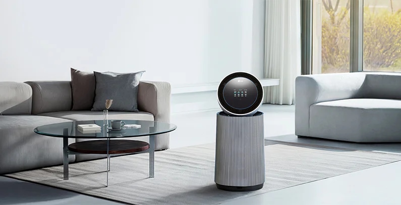 거실 공간에 LG 퓨리케어 AI 오브제컬렉션 360˚ 공기청정기가 놓여 있습니다. 양쪽에 베이지색 소파가 있으며, 쿠션과 담요가 놓여 있습니다. 유리 상판의 원형 테이블이 있고 그림과 검은색 스탠드 조명이 보입니다.