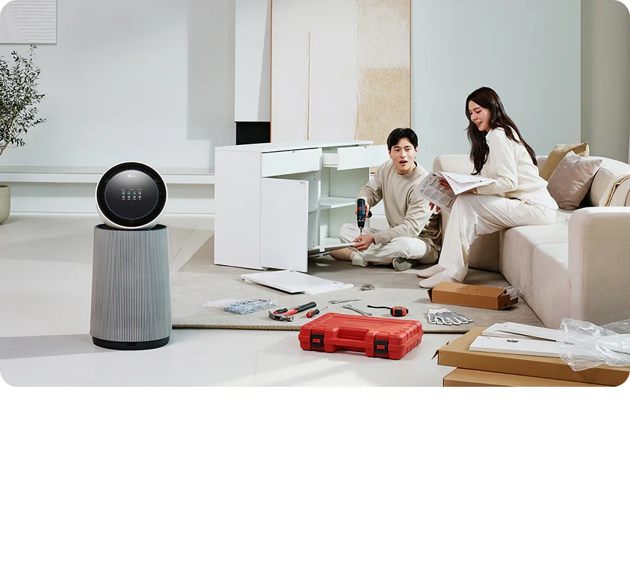 거실 공간에 LG 퓨리케어 AI 오브제컬렉션 360˚ 공기청정기가 놓여 있습니다. 가구를 조립하고 있는 두 사람의 모습이 보입니다. 주변에는 조립 중인 서랍장과 공구가 있습니다.