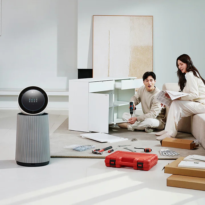 거실 공간에 LG 퓨리케어 AI 오브제컬렉션 360˚ 공기청정기가 놓여 있습니다. 가구를 조립하고 있는 두 사람의 모습이 보입니다. 주변에는 조립 중인 서랍장과 공구가 있습니다.