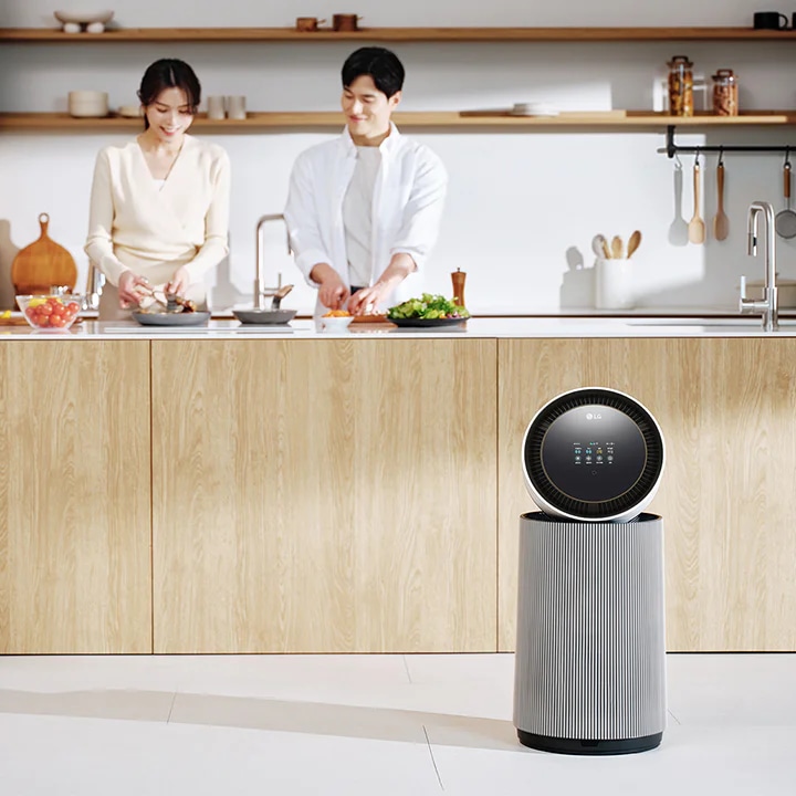 주방 공간에 LG 퓨리케어 AI 오브제컬렉션 360˚ 공기청정기가 있습니다. 나무 재질의 식탁과 의자가 놓여 있고 나무 장식의 주방 가구와 싱크대가 보입니다. 두 사람이 주방에서 요리를 준비하고 있는 모습이 함께 담겨 있습니다.