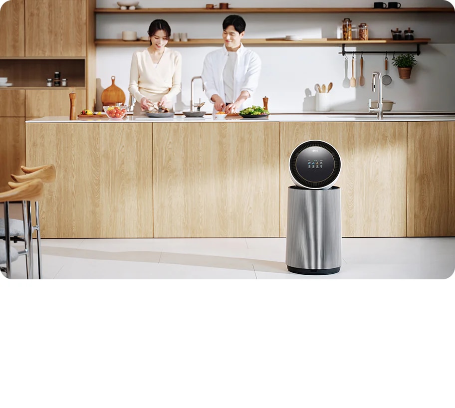 주방 공간에 LG 퓨리케어 AI 오브제컬렉션 360˚ 공기청정기가 있습니다. 나무 재질의 식탁과 의자가 놓여 있고 나무 장식의 주방 가구와 싱크대가 보입니다. 두 사람이 주방에서 요리를 준비하고 있는 모습이 함께 담겨 있습니다.