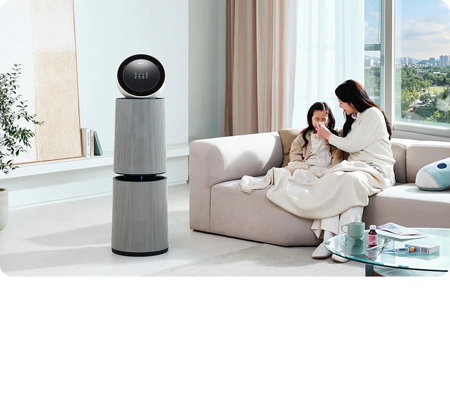 거실 공간에 LG 퓨리케어 AI 오브제컬렉션 360˚ 공기청정기가 놓여 있습니다. 소파 위에는 두 사람이 담요를 덮고 앉아 있으며, 한 여성이 아이의 코를 풀어주고 있는 모습입니다. 테이블 위에는 컵, 약봉투, 약이 놓여 있습니다.